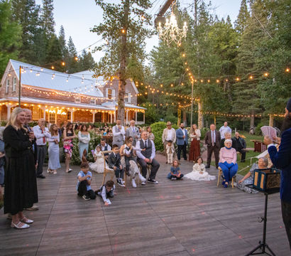 Evening outdoor wedding reception at a rustic farmhouse surrounded by trees, string lights and a hanging chandelier, guests gathered on a wooden dance floor watching a performer with playing cards.