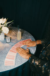 Hands fanning red-backed playing cards across a blue velvet tablecloth next to a champagne-style cocktail with a star stirrer and a silver vase of white roses — intimate lounge card trick scene.