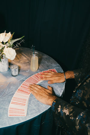 Hands fanning red-backed playing cards across a blue velvet tablecloth next to a champagne-style cocktail with a star stirrer and a silver vase of white roses — intimate lounge card trick scene.