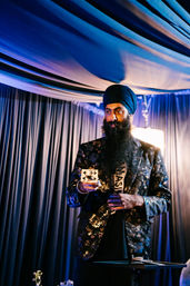 Performer wearing a turban and patterned blazer doing a card trick on an indoor stage with blue drapery and dramatic lighting