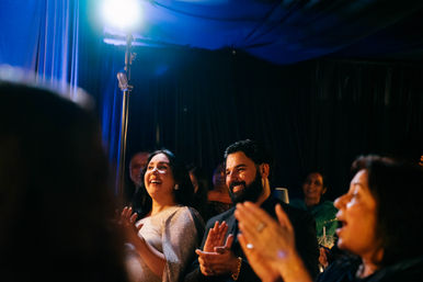 Smiling couple — woman in a sparkly dress and a bearded man — and guests applauding at an indoor evening celebration under blue stage lighting and a bright spotlight.