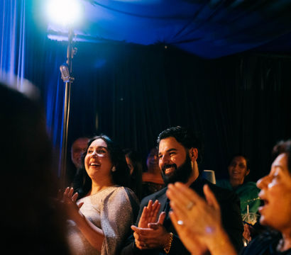 Smiling couple — woman in a sparkly dress and a bearded man — and guests applauding at an indoor evening celebration under blue stage lighting and a bright spotlight.