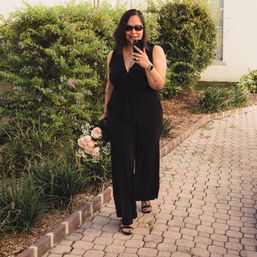 Woman in a black jumpsuit and sunglasses walking down a brick‑paved garden path, holding a bouquet of pale pink roses and baby’s breath while smiling and taking a selfie.