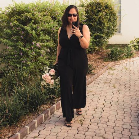 Woman in a black jumpsuit and sunglasses walking down a brick‑paved garden path, holding a bouquet of pale pink roses and baby’s breath while smiling and taking a selfie.