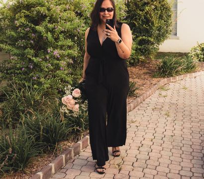 Woman in a black jumpsuit and sunglasses walking down a brick‑paved garden path, holding a bouquet of pale pink roses and baby’s breath while smiling and taking a selfie.