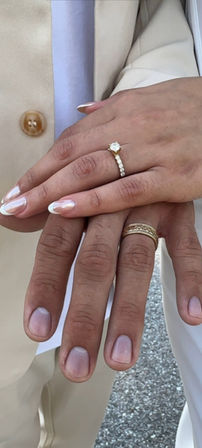 Close-up of stacked bride and groom hands showing a solitaire diamond engagement ring with a diamond band and a textured gold wedding band, bride’s French-manicured nails and cream suit sleeve visible