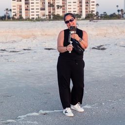 Smiling woman on a sandy beach at sunset filming with a smartphone gimbal, wearing sunglasses, black outfit and white sneakers, coastal condos and palm trees in the background