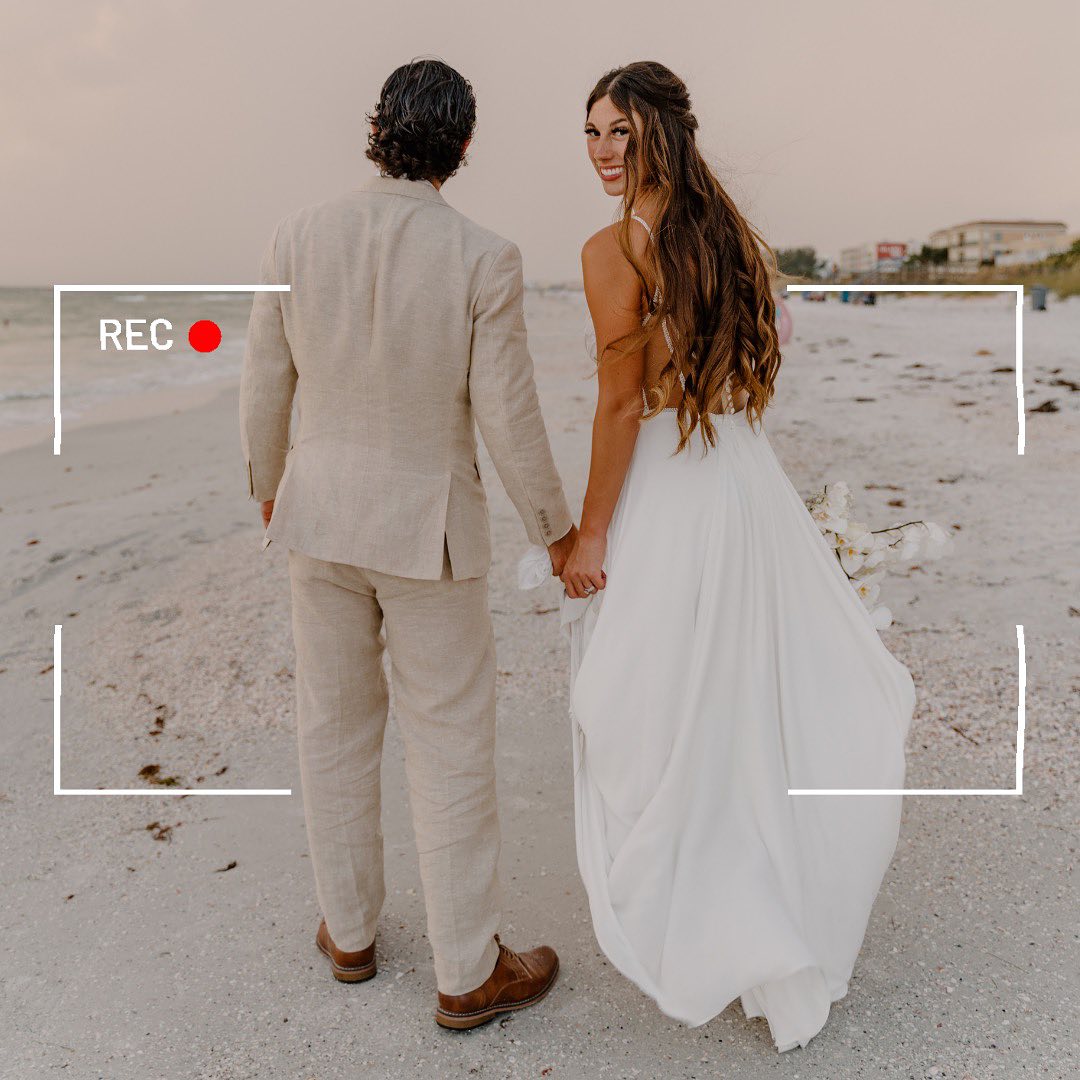 Bride in flowing white gown and groom in a light linen suit holding hands as they walk along a sandy beach shoreline at sunset with ocean waves and a faux video REC overlay