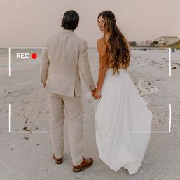 Bride in flowing white gown and groom in a light linen suit holding hands as they walk along a sandy beach shoreline at sunset with ocean waves and a faux video REC overlay