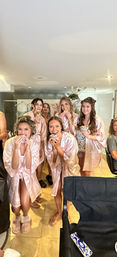 Bridal party in a bright dressing suite wearing matching pink satin robes, smiling and holding wrapped sandwiches while getting ready together.