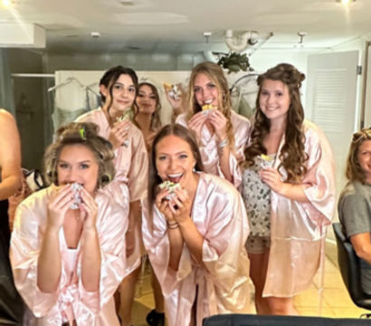 Bridal party in a bright dressing suite wearing matching pink satin robes, smiling and holding wrapped sandwiches while getting ready together.