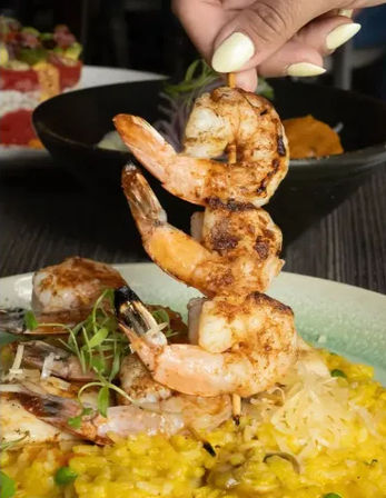 Charred grilled shrimp skewers lifted above a plate of creamy saffron risotto with peas, microgreens and shaved cheese — vibrant seafood restaurant dish.