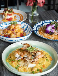 Colorful seafood spread on a gray wood table at a restaurant: grilled shrimp skewers over creamy saffron risotto with peas and carrots, seared scallops in patterned bowls, a floral-garnished plate and vase of flowers.
