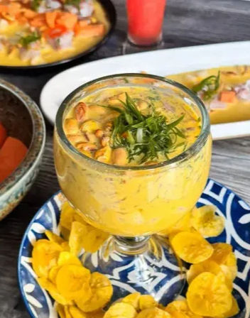 Peruvian-style seafood ceviche in a goblet with creamy yellow sauce, toasted corn and herbs, served with plantain chips on a blue patterned plate on a rustic wooden table
