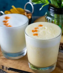 Two frothy citrus cocktails in short glasses topped with orange zest, served on a wooden board with jarred herbs and snacks in the background
