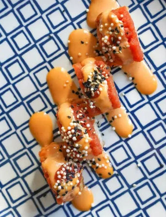 Three sushi roll pieces topped with spicy orange mayo, black-and-white sesame seeds and red fish, arranged on a blue geometric patterned plate.