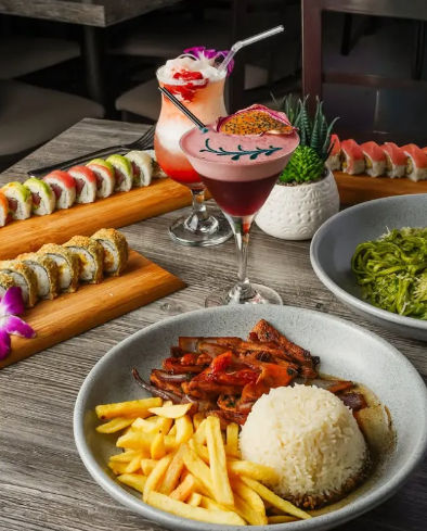 Vibrant Asian-fusion spread with colorful sushi rolls on wooden boards, two tropical cocktails garnished with flowers and dragon fruit, and a bowl of rice, fries and stir-fried meat on a rustic table