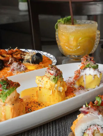 Peruvian-inspired causa bites topped with avocado, spicy mayo and crab on a white platter, sushi pieces nearby and a mango cocktail in a tiki glass