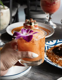 Hand holding a peach-orange cocktail rimmed with chili salt and topped with a purple orchid, on a restaurant table with colorful plates and drinks in the background.