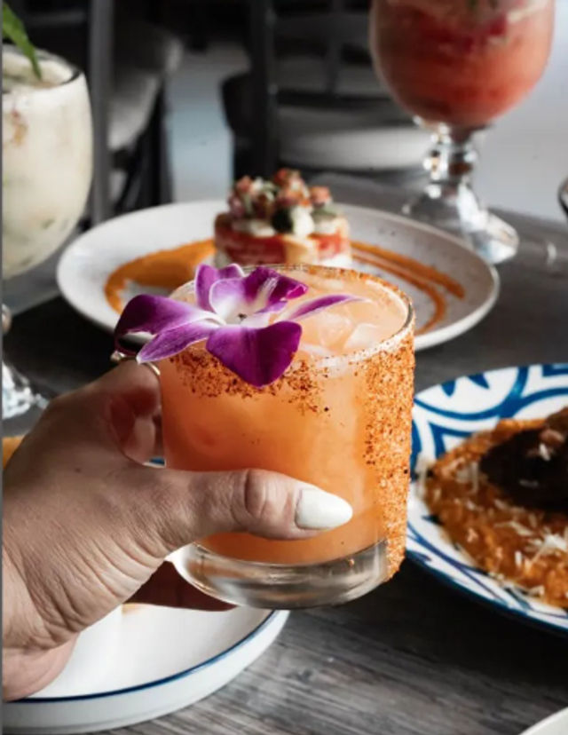 Hand holding a peach-orange cocktail rimmed with chili salt and topped with a purple orchid, on a restaurant table with colorful plates and drinks in the background.