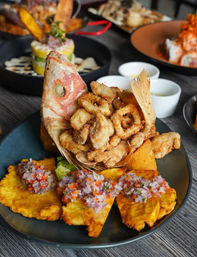 Crispy fried seafood piled inside a fried fish head, served with golden plantain tostones topped with pico de gallo and small dipping sauces on a dark plate — Latin American coastal seafood platter.