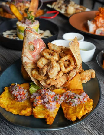 Crispy fried seafood piled inside a fried fish head, served with golden plantain tostones topped with pico de gallo and small dipping sauces on a dark plate — Latin American coastal seafood platter.