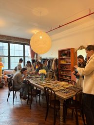 Sunlit urban loft boutique with a large round paper lantern over a wooden table covered in jewelry, watches and trays, clothing racks and shelves in the background and shoppers browsing accessories.