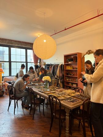 Sunlit urban loft boutique with a large round paper lantern over a wooden table covered in jewelry, watches and trays, clothing racks and shelves in the background and shoppers browsing accessories.