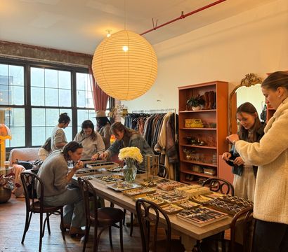 Sunlit urban loft boutique with a large round paper lantern over a wooden table covered in jewelry, watches and trays, clothing racks and shelves in the background and shoppers browsing accessories.