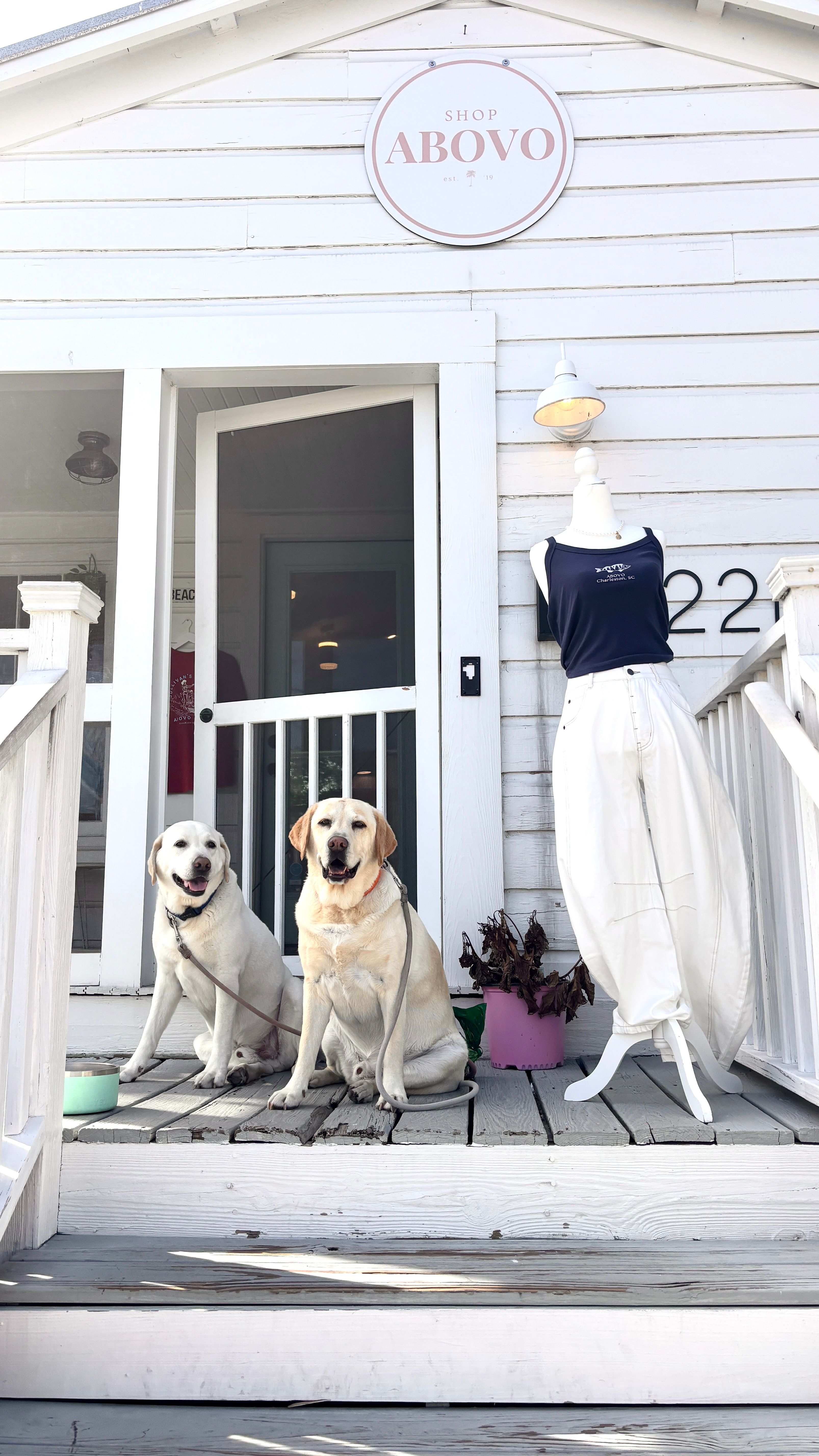 Two yellow Labradors sitting on a weathered white wooden boutique porch beside a mannequin in white pants and navy tank, round shop sign above the entrance