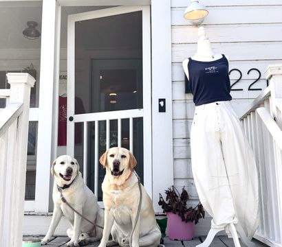 Two yellow Labradors sitting on a weathered white wooden boutique porch beside a mannequin in white pants and navy tank, round shop sign above the entrance