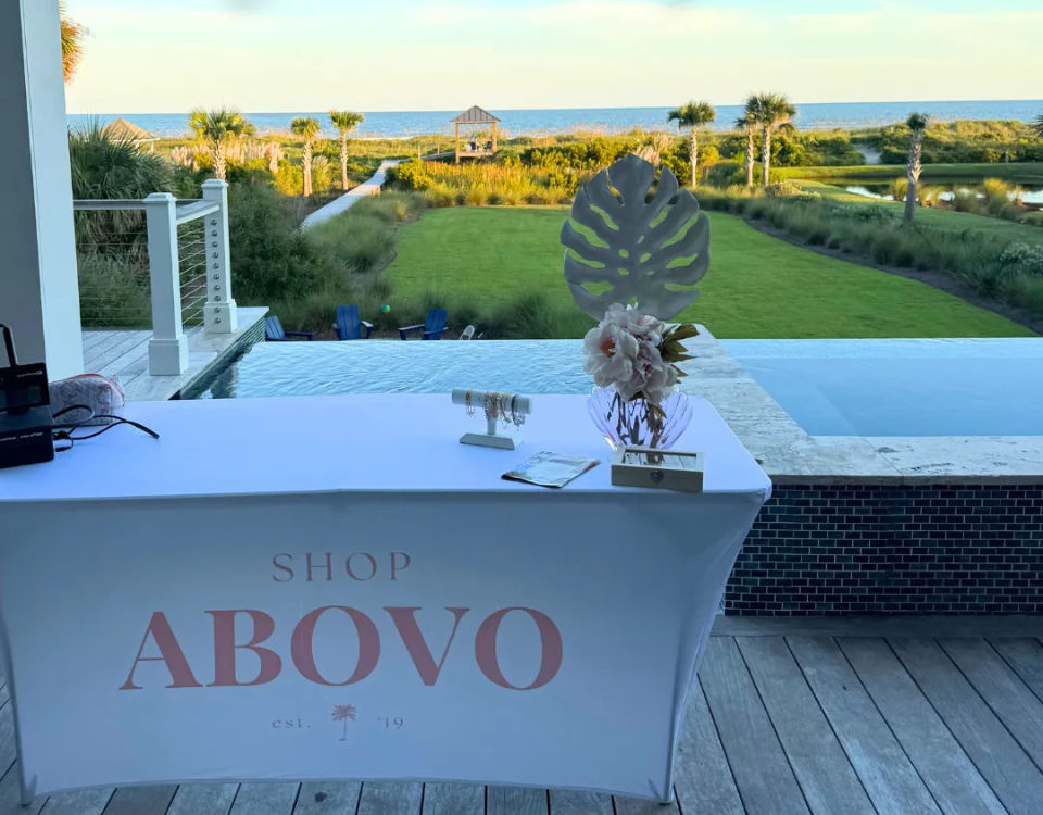 Oceanfront vendor table draped in a branded cloth with a vase of flowers and small jewelry display by an infinity pool, overlooking a palm‑lined lawn, sand dunes and the sea.