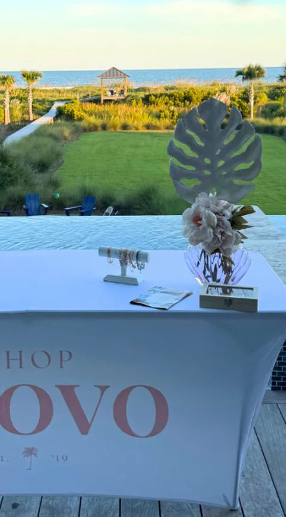 Oceanfront vendor table draped in a branded cloth with a vase of flowers and small jewelry display by an infinity pool, overlooking a palm‑lined lawn, sand dunes and the sea.