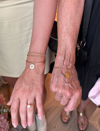 Close-up of a younger and older hand side-by-side wearing gold chain bracelets with sun and disc charms and a dainty gold solitaire ring — jewelry lifestyle close-up.