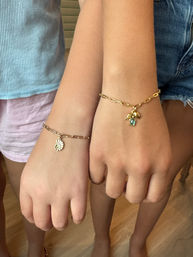 Close-up of two children's wrists side-by-side wearing gold chain charm bracelets — one with a nautilus shell charm, the other with a blue gemstone and leaf charm, paired with casual summer clothing.