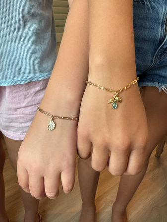 Close-up of two children's wrists side-by-side wearing gold chain charm bracelets — one with a nautilus shell charm, the other with a blue gemstone and leaf charm, paired with casual summer clothing.