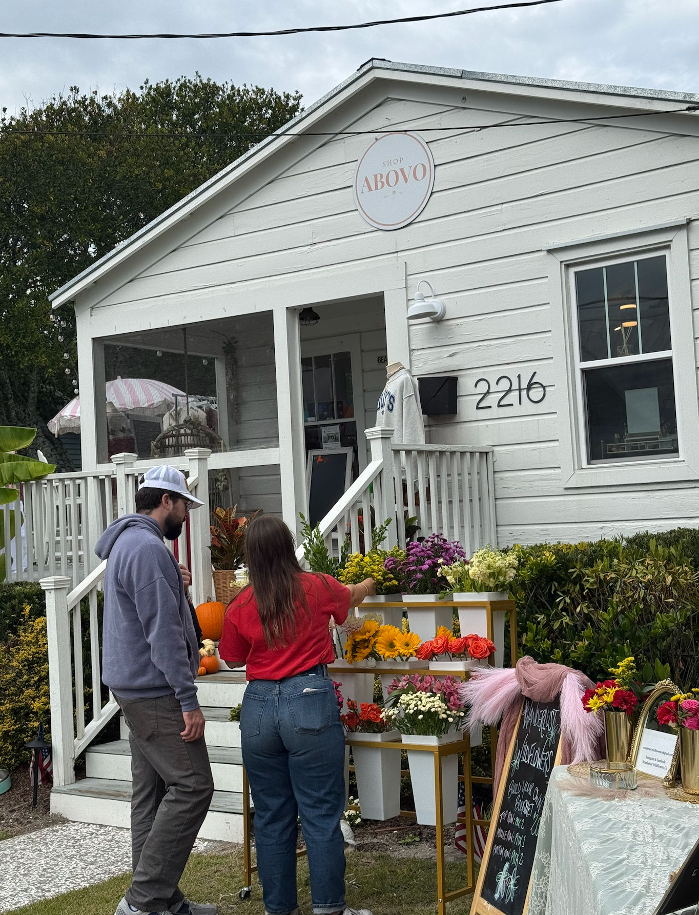 White cottage-style boutique with front porch and outdoor flower stand — sunflowers, roses, mums and pumpkins — two shoppers choosing bouquets