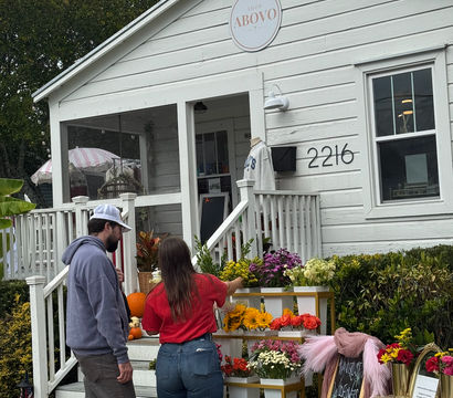 White cottage-style boutique with front porch and outdoor flower stand — sunflowers, roses, mums and pumpkins — two shoppers choosing bouquets