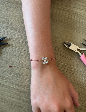 Close-up of a wrist wearing a handmade delicate gold chain bracelet with a crystal flower charm and pink ruby beads, jewelry pliers and a loose bead on a wooden tabletop — DIY jewelry scene.