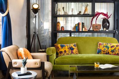 Eclectic urban living room with a green velvet sofa and yellow floral pillows, beige armchair with orange cushion, tripod floor lamp, black shelving with books and decor, and coffee table with a drink and open book