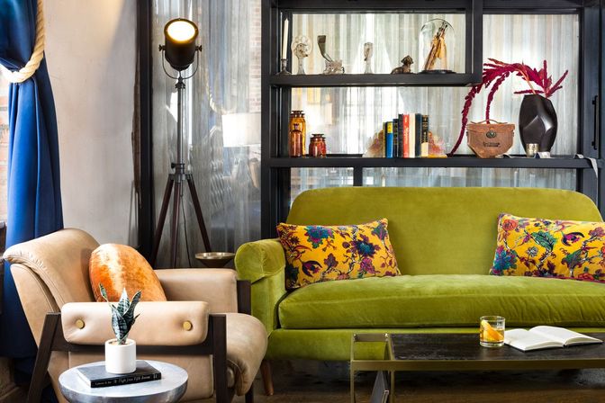 Eclectic urban living room with a green velvet sofa and yellow floral pillows, beige armchair with orange cushion, tripod floor lamp, black shelving with books and decor, and coffee table with a drink and open book