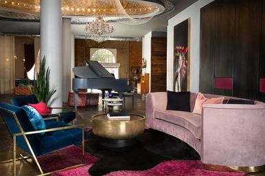 Boutique hotel lounge with a pink curved velvet sofa, blue velvet armchairs with brass frames, round brass coffee table, grand piano under a crystal chandelier, colorful artwork and plants.