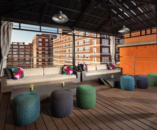 Rooftop urban lounge with shaded metal canopy, beige sectional sofas, colorful throw pillows, knitted poufs and low coffee tables on wood decking overlooking red-brick apartment buildings.