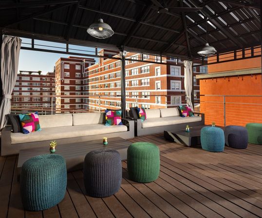 Rooftop urban lounge with shaded metal canopy, beige sectional sofas, colorful throw pillows, knitted poufs and low coffee tables on wood decking overlooking red-brick apartment buildings.