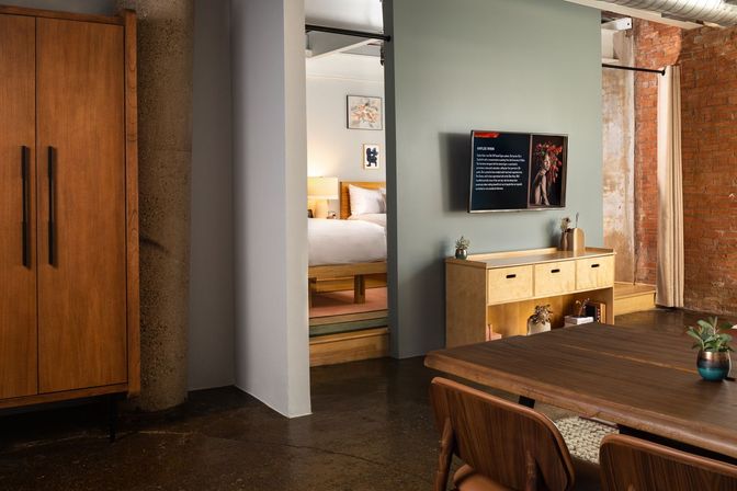 Cozy industrial loft living area with exposed brick wall, open-plan dining table and chairs, mid-century wooden sideboard with wall TV, and a bedroom alcove with a platform bed.