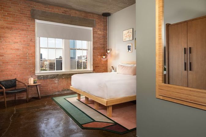 Industrial-style downtown loft bedroom with exposed brick wall, large city-view window, minimalist wooden platform bed and geometric rug