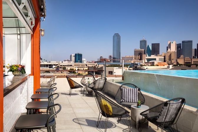 Rooftop pool terrace with bar stools, modern wire-frame lounge chairs and small tables, colorful cushions and a planter, overlooking a downtown skyline of glass skyscrapers under a clear blue sky.