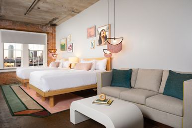 Industrial-chic loft-style hotel room with exposed brick and concrete ceiling, two beds with white linens, pink-fringe pendant lights and framed art, gray sofa with teal pillows, white modern coffee table on a geometric rug, and a large window showing a downtown skyline view.