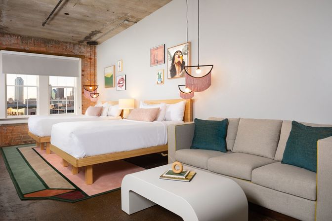 Industrial-chic loft-style hotel room with exposed brick and concrete ceiling, two beds with white linens, pink-fringe pendant lights and framed art, gray sofa with teal pillows, white modern coffee table on a geometric rug, and a large window showing a downtown skyline view.