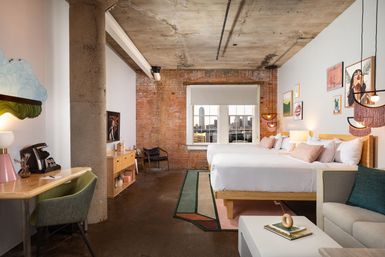Sunlit industrial-chic loft hotel room with exposed brick and concrete ceiling, two queen beds with pastel pillows, eclectic wall art, desk, sofa and window framing a downtown urban skyline